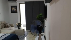 Un dormitorio moderno con cama, ventilador de pie, televisor montado en la pared y una puerta que da a otra habitación.