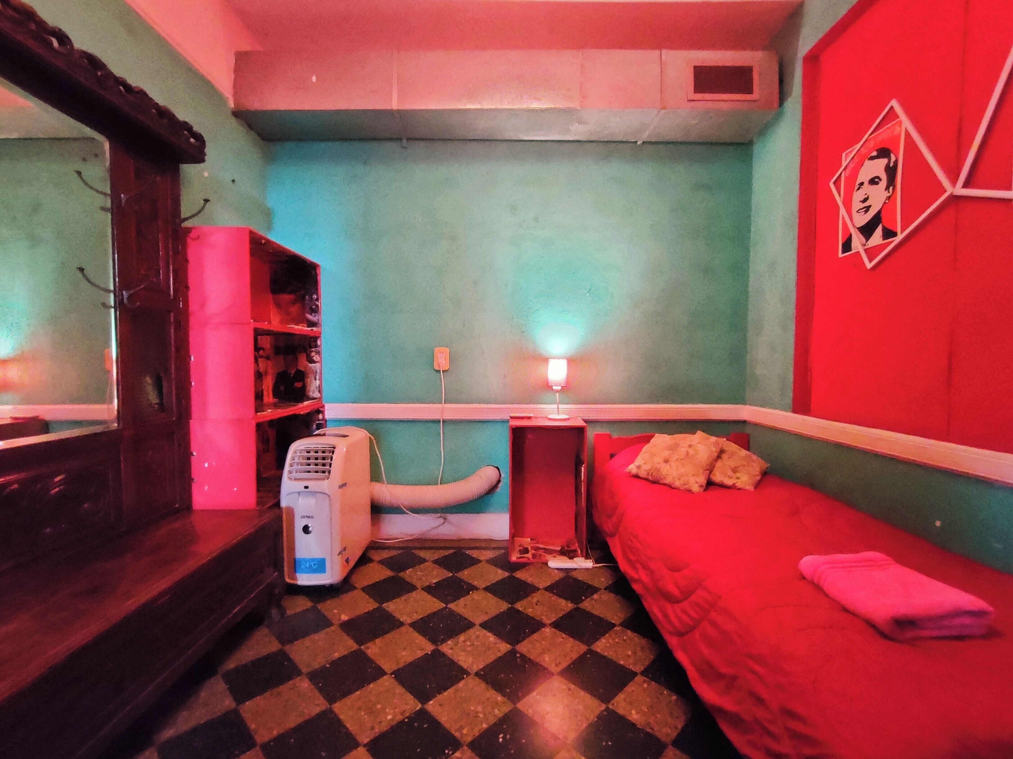 A room with a checkered floor, a red bed, a desk with a lamp, and a heater.