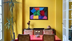 Uma área de jantar com uma mesa e cadeiras vermelhas, um ventilador de teto, uma planta grande e uma pintura abstrata colorida na parede.