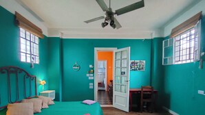 Quarto com paredes verde-água, uma cama com almofadas, uma escrivaninha e um ventilador de teto.