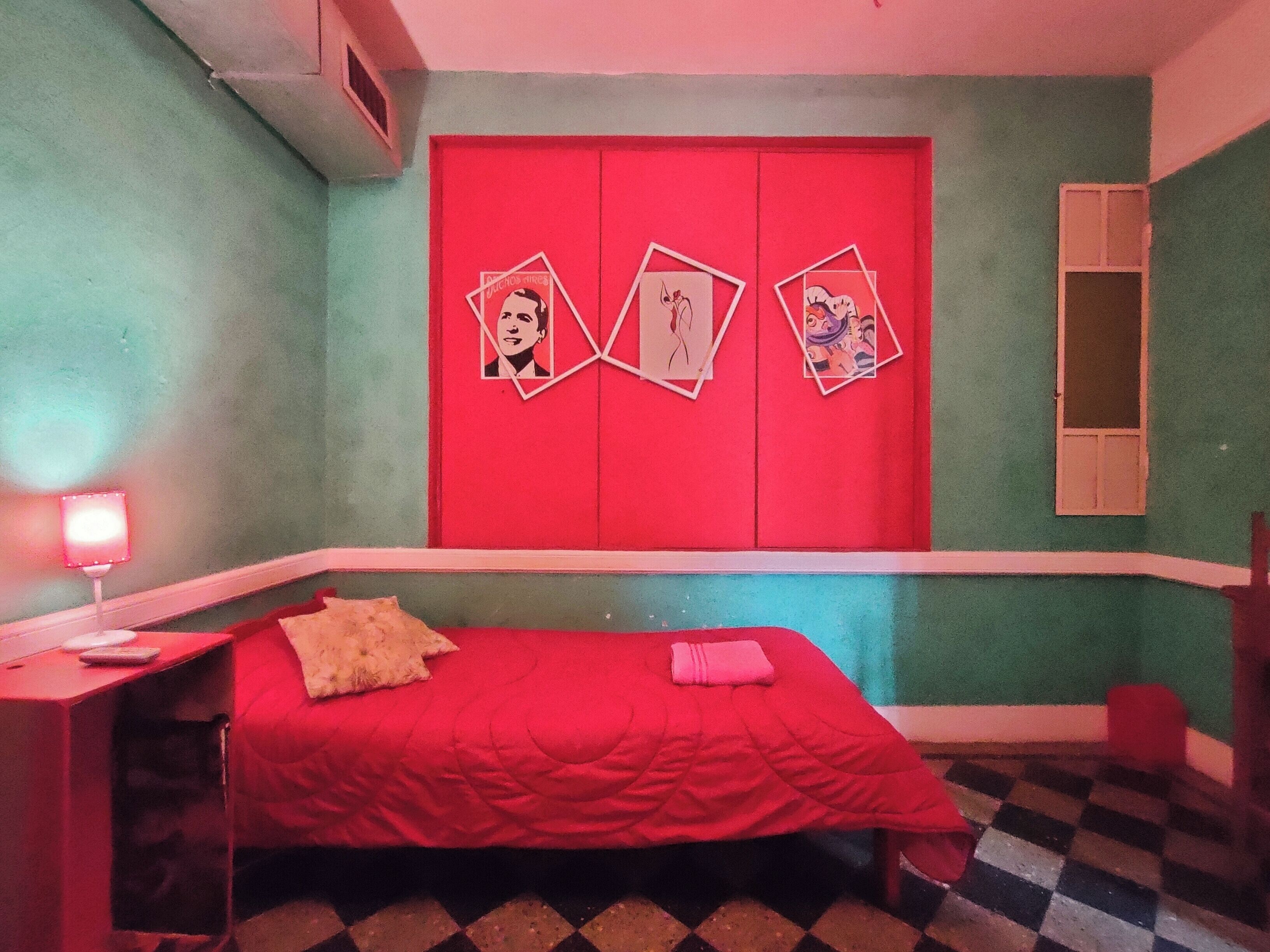A bedroom with a red bed, a checkered floor, and a red wall with three framed pictures.