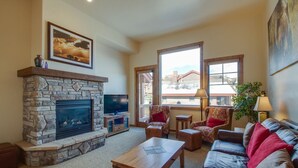 A cozy living room with a stone fireplace, comfortable seating, and large windows offering a view of the outside.