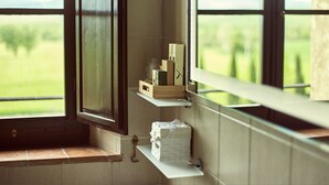 Un baño con ventana que da a un campo verde y montañas. Hay dos estantes con artículos de aseo y un pequeño objeto blanco con diseño de ladrillos.