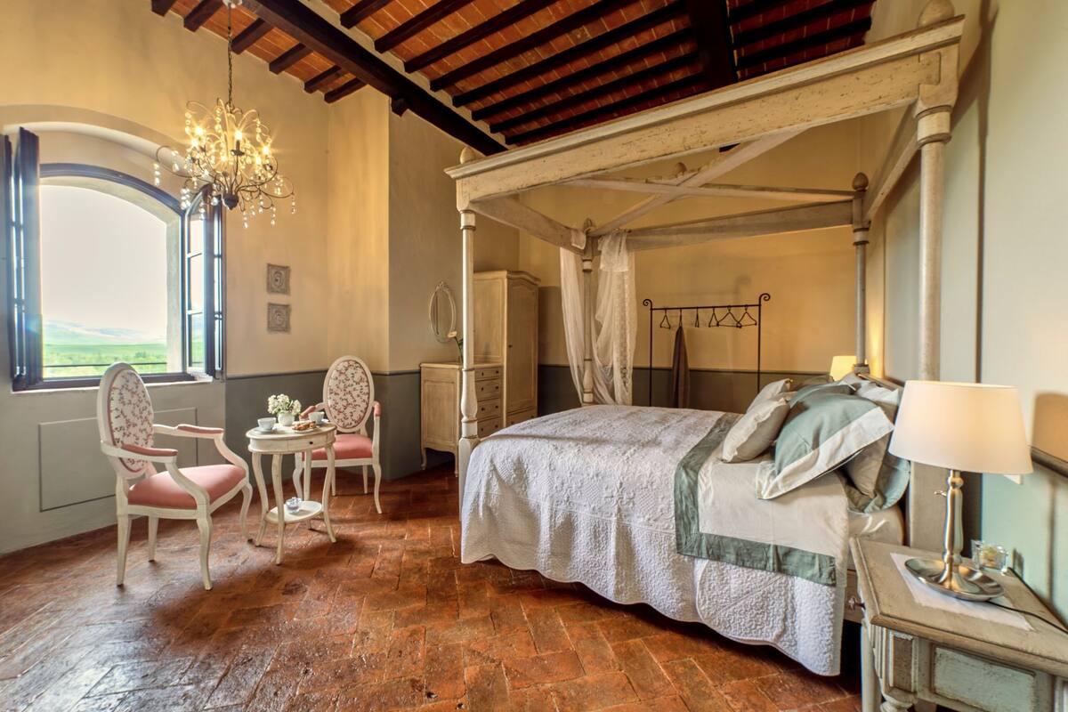 Une chambre à coucher avec un lit à baldaquin, un lustre, une fenêtre donnant sur un beau paysage, un fauteuil et une petite table.