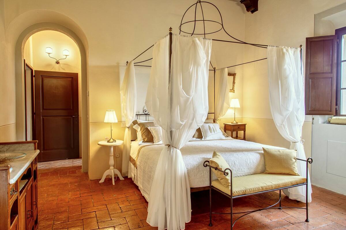 Un lit à baldaquin à quatre poteaux, agrémenté de draps blancs, une table de chevet avec une lampe et un petit fauteuil avec des coussins.