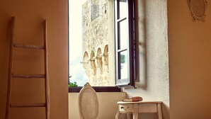 Una habitación con una escalera de madera, una silla de mimbre, una mesita con un libro y un par de zapatos, y una ventana que da a un edificio de piedra.