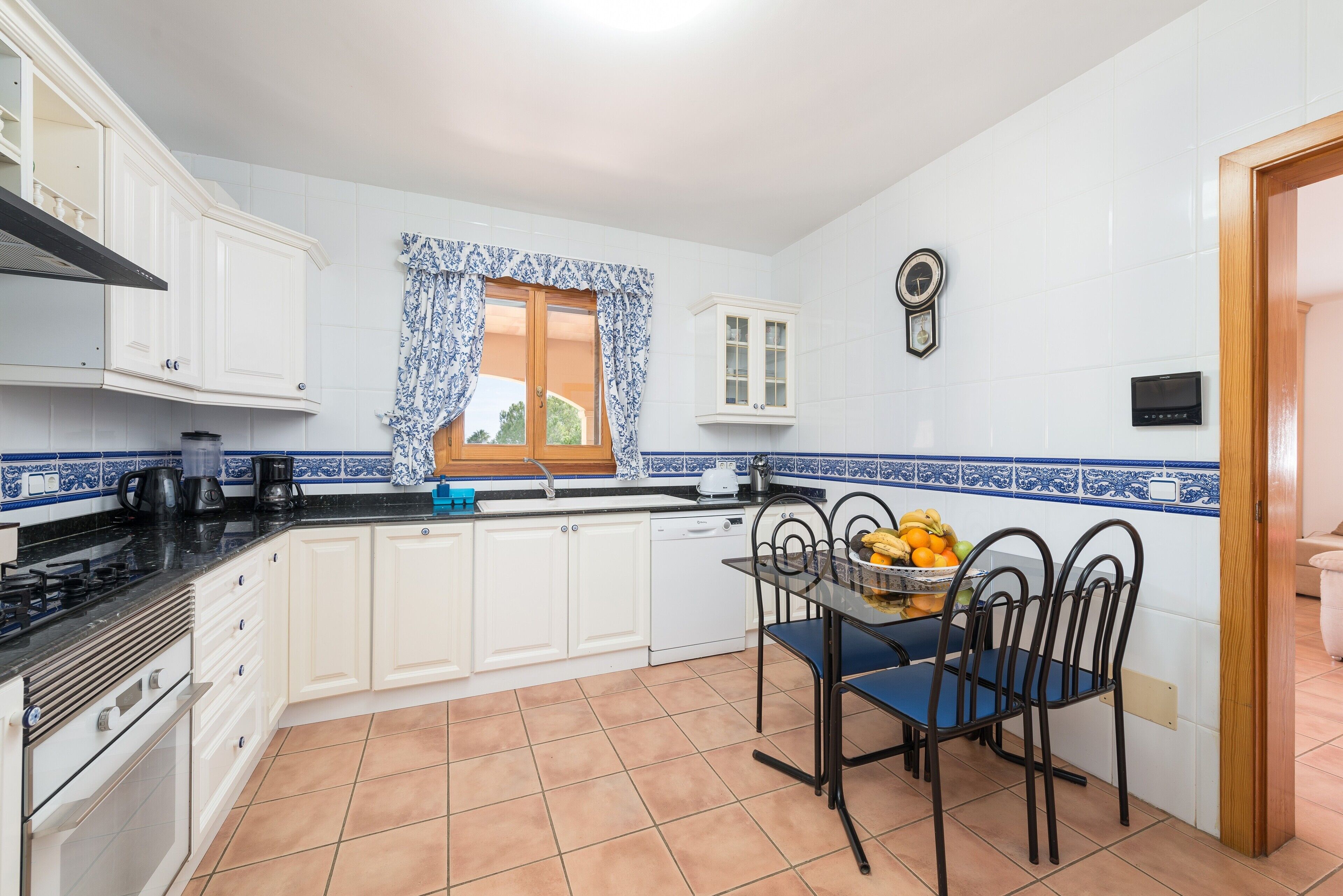 Una cocina con armarios blancos, encimera negra, ventana con cortinas azules, un reloj en la pared y un frutero sobre la mesa.