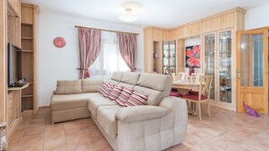 Ein Wohnzimmer mit einem beigefarbenen Ecksofa, einem Esstisch mit Stühlen, Holzschränken und einem großen Fenster mit Vorhängen.