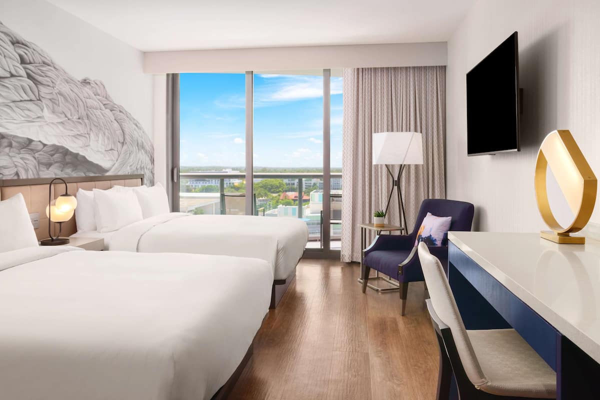 Een hotelkamer met twee bedden, een bureau, een stoel, een televisie en een groot raam met uitzicht op de stad.