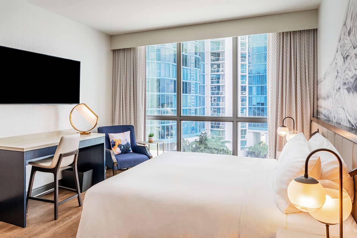 Een hotelkamer met een groot bed, een bureau, een stoel en uitzicht op hoge gebouwen door grote ramen.