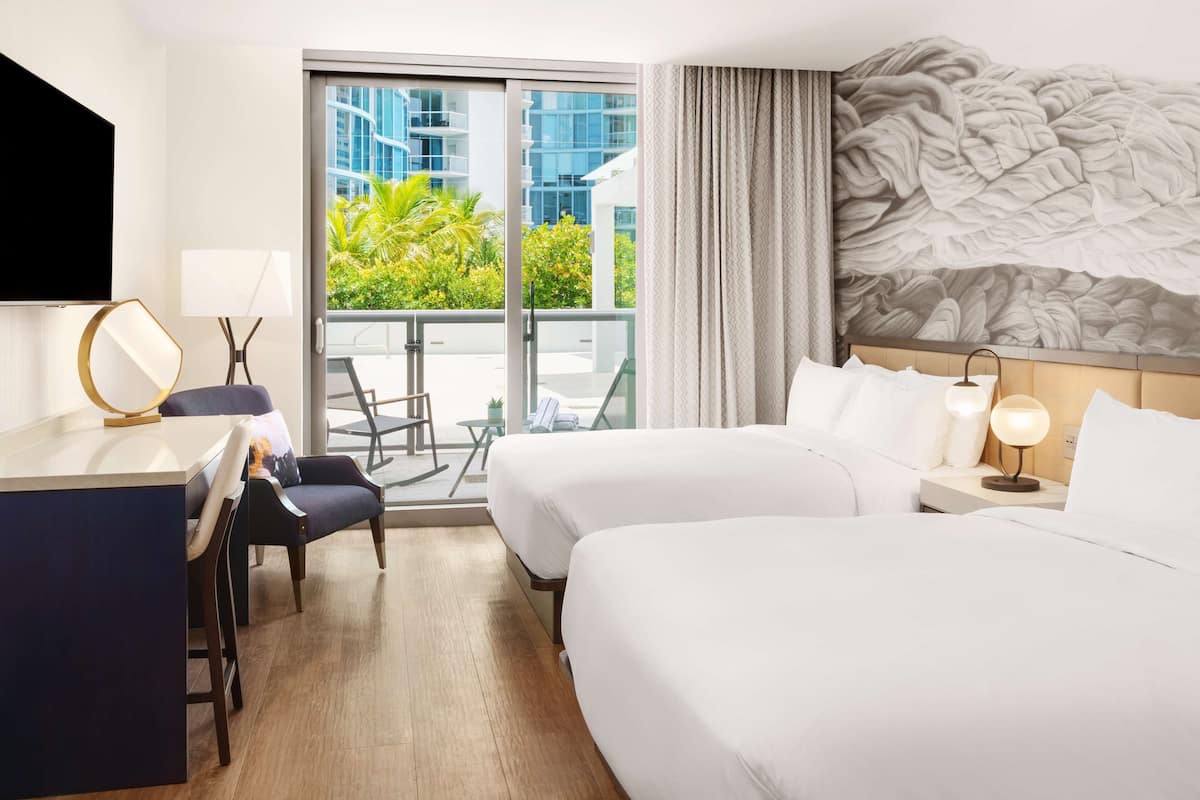 Hotelkamer met een groot bed, een bureau, een stoel en een balkon met uitzicht op groen en gebouwen.