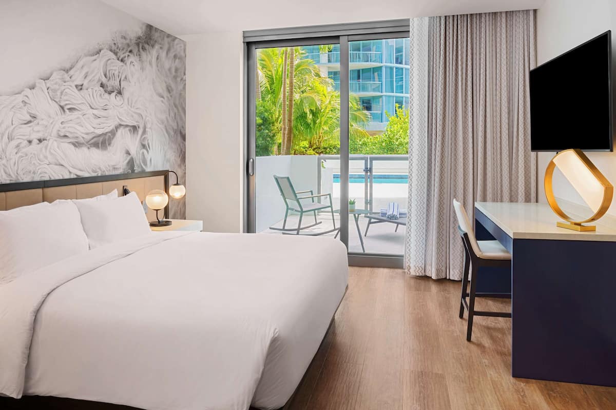 Hotelkamer met een groot bed, een bureau met stoel, een televisie en een balkon met uitzicht op groen en gebouwen.