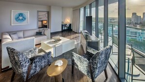 A modern hotel room with a large balcony, a sofa, armchairs, a coffee table, and a desk with a lamp.