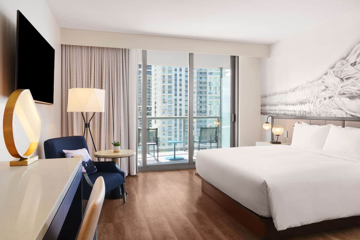 Hotelkamer met een bed, een bureau, een stoel, een televisie, een lamp, een bijzettafeltje met een plant en uitzicht op hoogbouw door grote ramen.