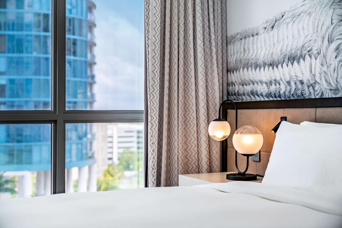 Een hotelkamer met een groot raam, een bed, twee bedlampjes en een kunstwerk aan de muur.
