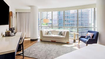 A modern hotel room with a large window, a sofa, a chair, a small table, and a bed.