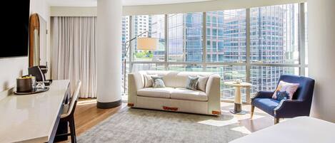 A modern hotel room with a large window, a sofa, a chair, a small table, and a bed.