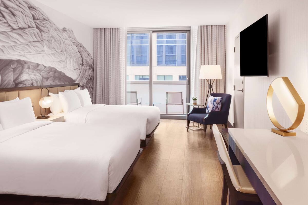 Hotelkamer met twee bedden, een televisie, een bureau met stoel, een groot raam en uitzicht op de stad.