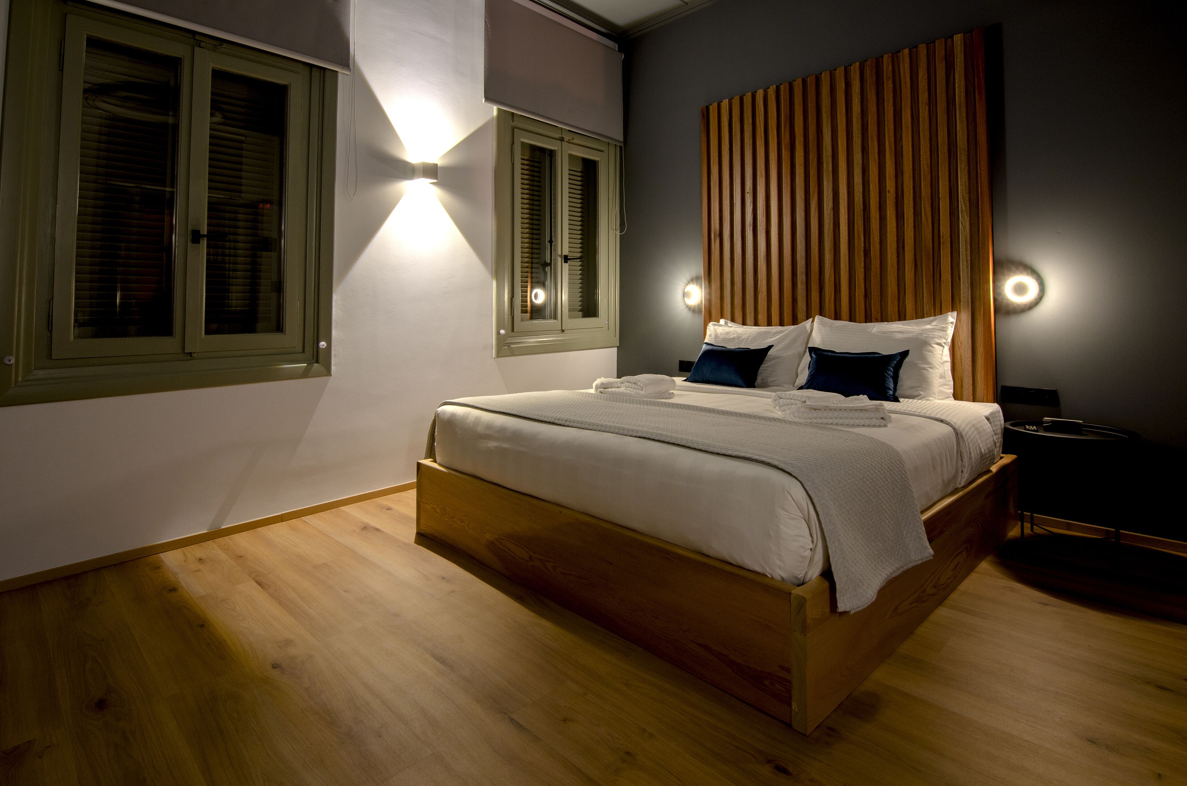 A bedroom with a wooden bed, two windows with shutters, and wall-mounted lighting.