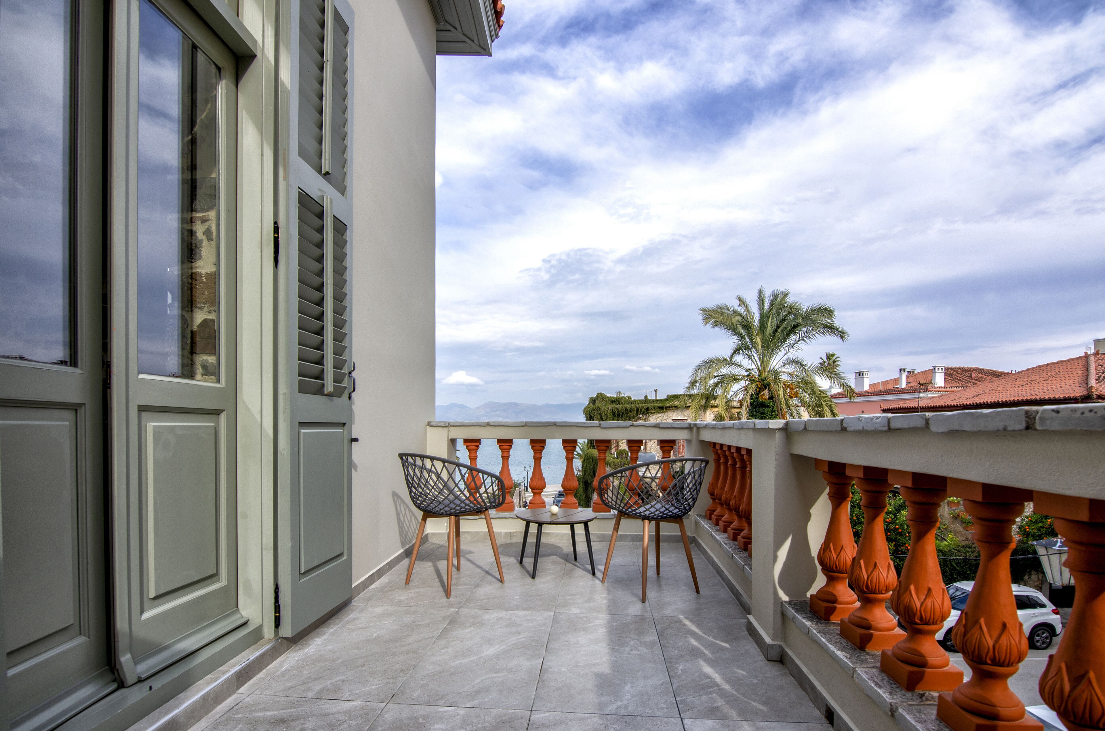 A balcony with two chairs and a small table, overlooking a cityscape with palm trees and buildings.