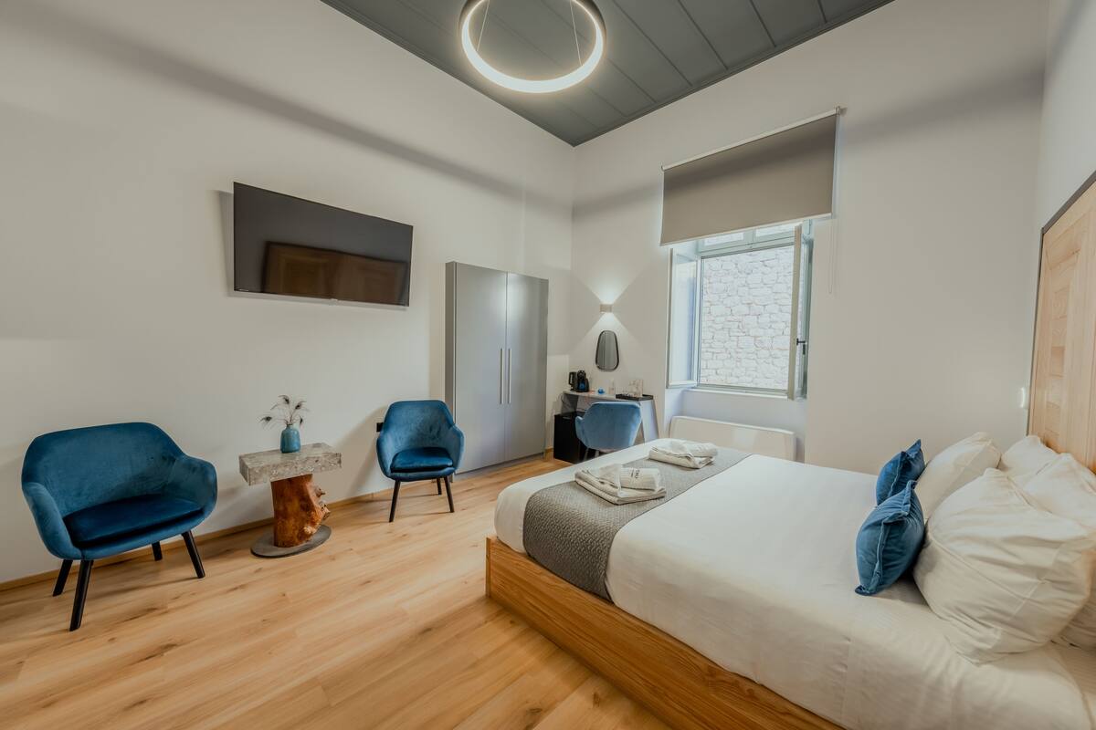 Une chambre à coucher moderne avec un grand lit, deux fauteuils bleus, une table basse en bois et un bureau avec une lampe.