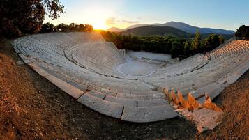 Un anfiteatro antiguo con asientos escalonados y un escenario central, rodeado de árboles y montañas al fondo.