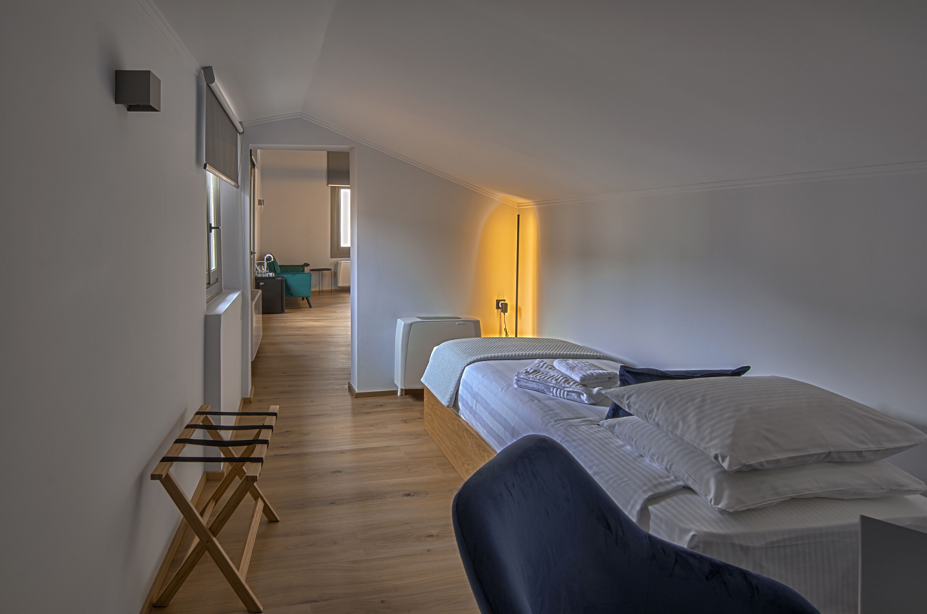 Un dormitorio moderno con suelo de madera, una cama con ropa blanca, una mesita de noche y una mesilla auxiliar. Se ve una ventana en el lado izquierdo y una puerta que da a otra habitación.
