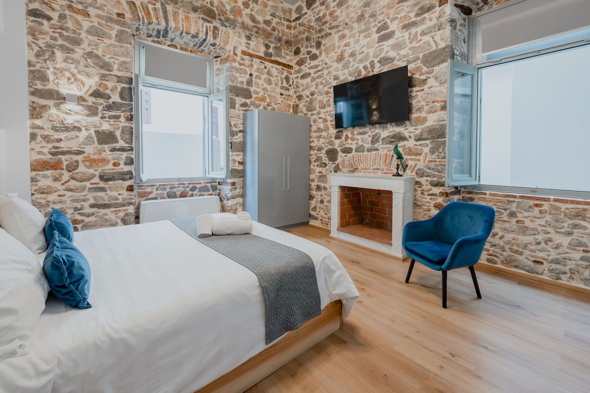 Une chambre avec un mur en pierre, un lit avec une literie blanche, une chaise bleue et une cheminée.