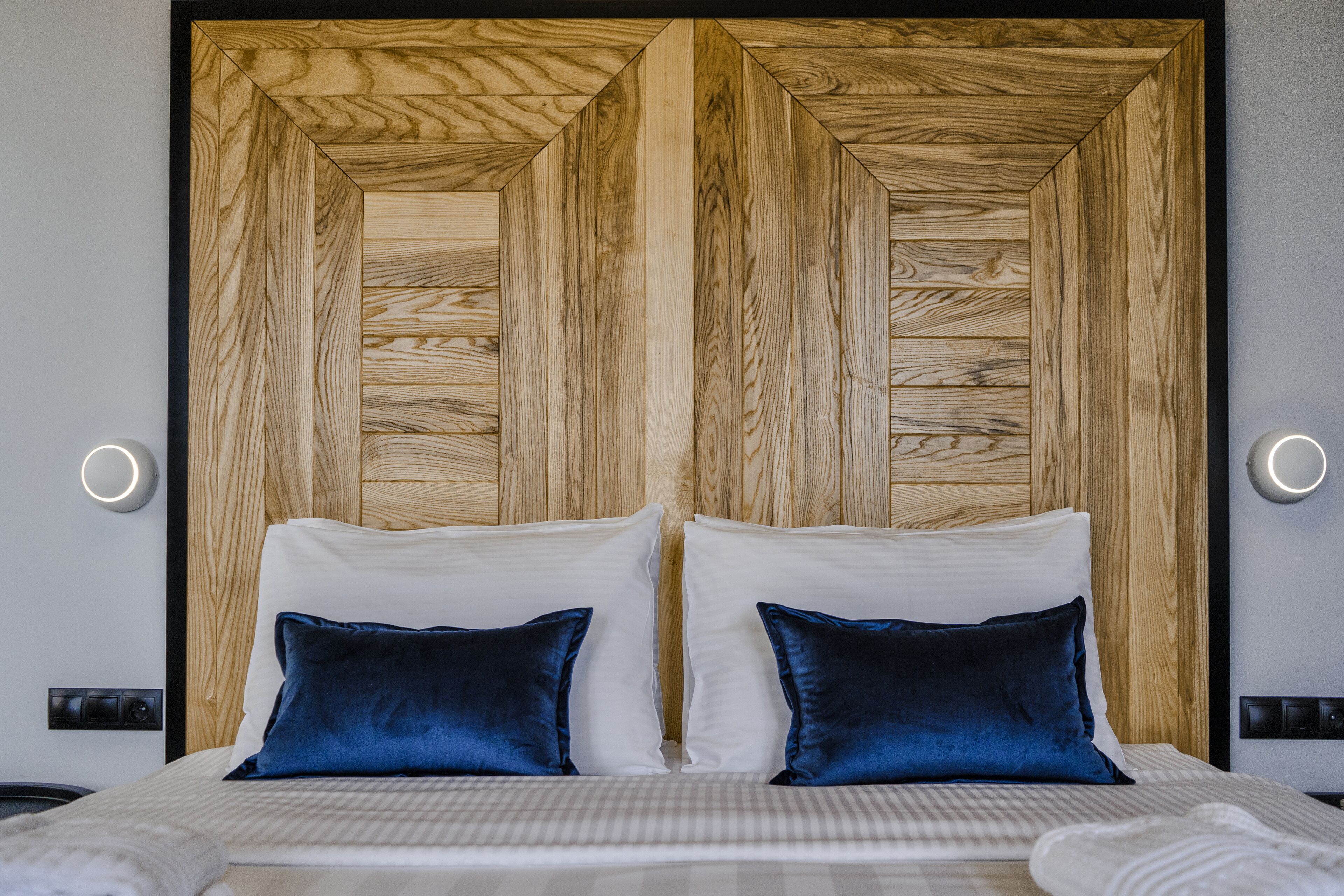 A double bed with white and dark blue pillows, a wooden headboard, and two wall-mounted lights.
