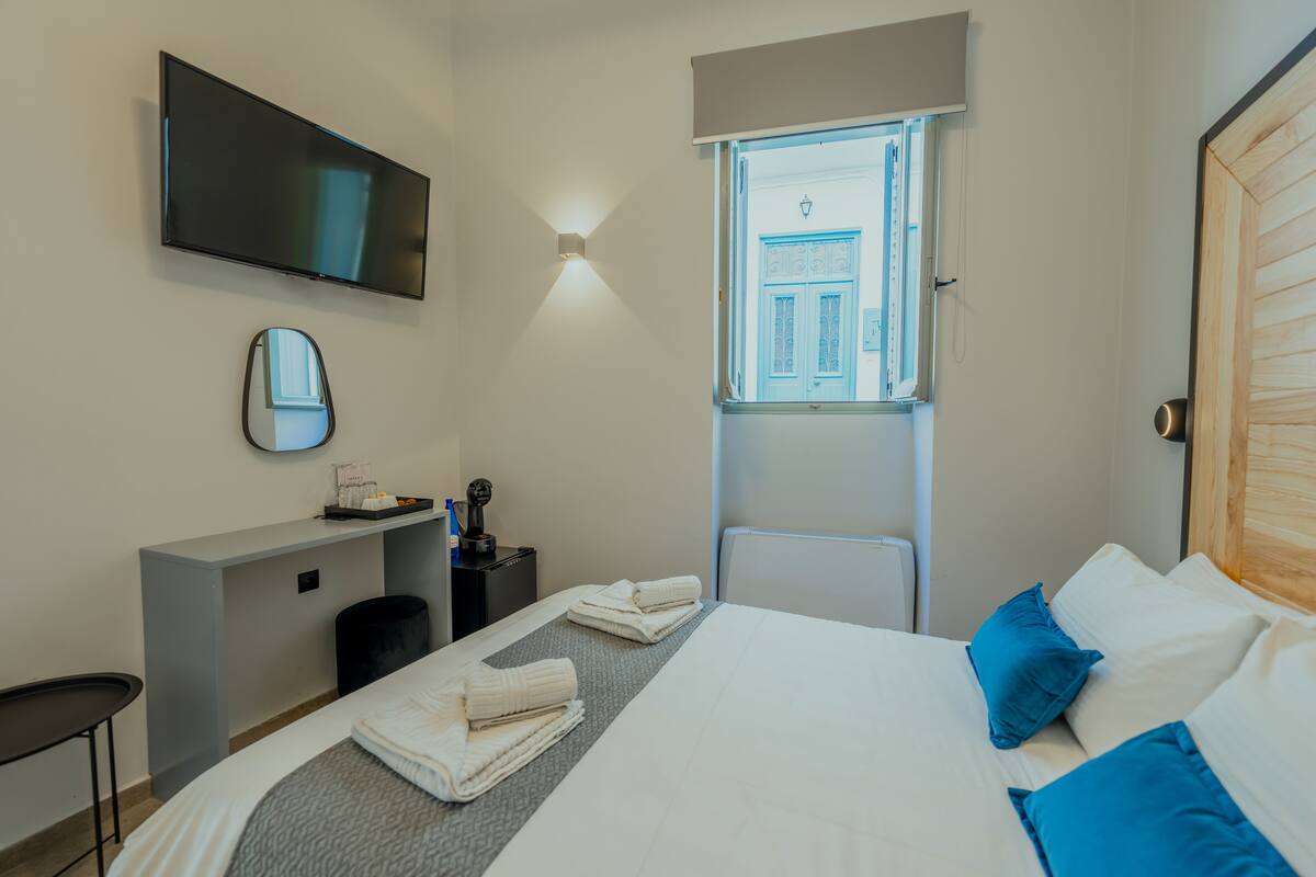 Une chambre d’hôtel équipée d’un lit, d’un bureau, d’une télévision et d’une fenêtre donnant sur un immeuble.