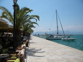 Un café frente al mar con terraza, una fila de palmeras y barcos al fondo.