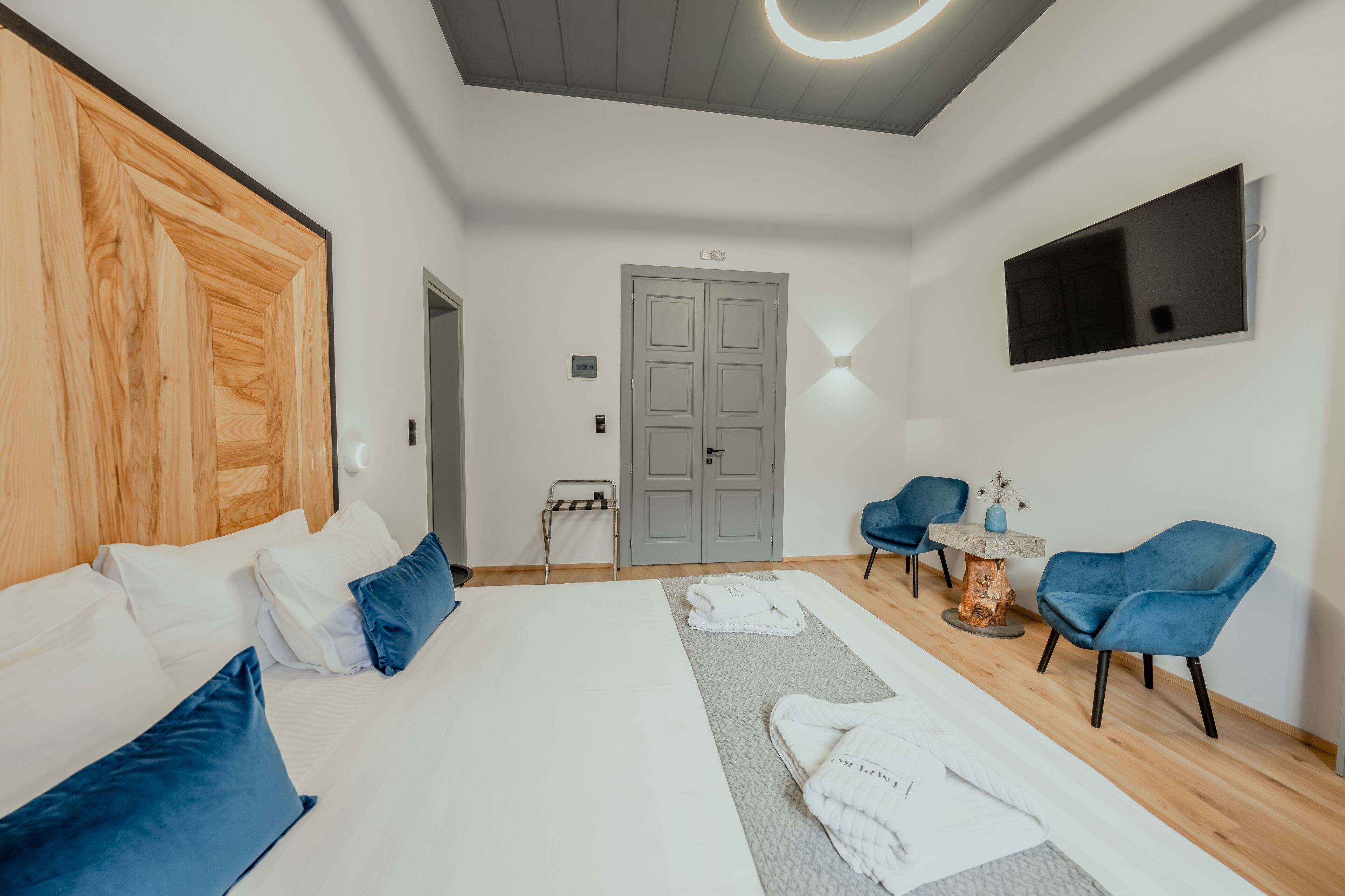 A modern hotel room with a large bed, two blue chairs, a TV, and a wooden headboard.