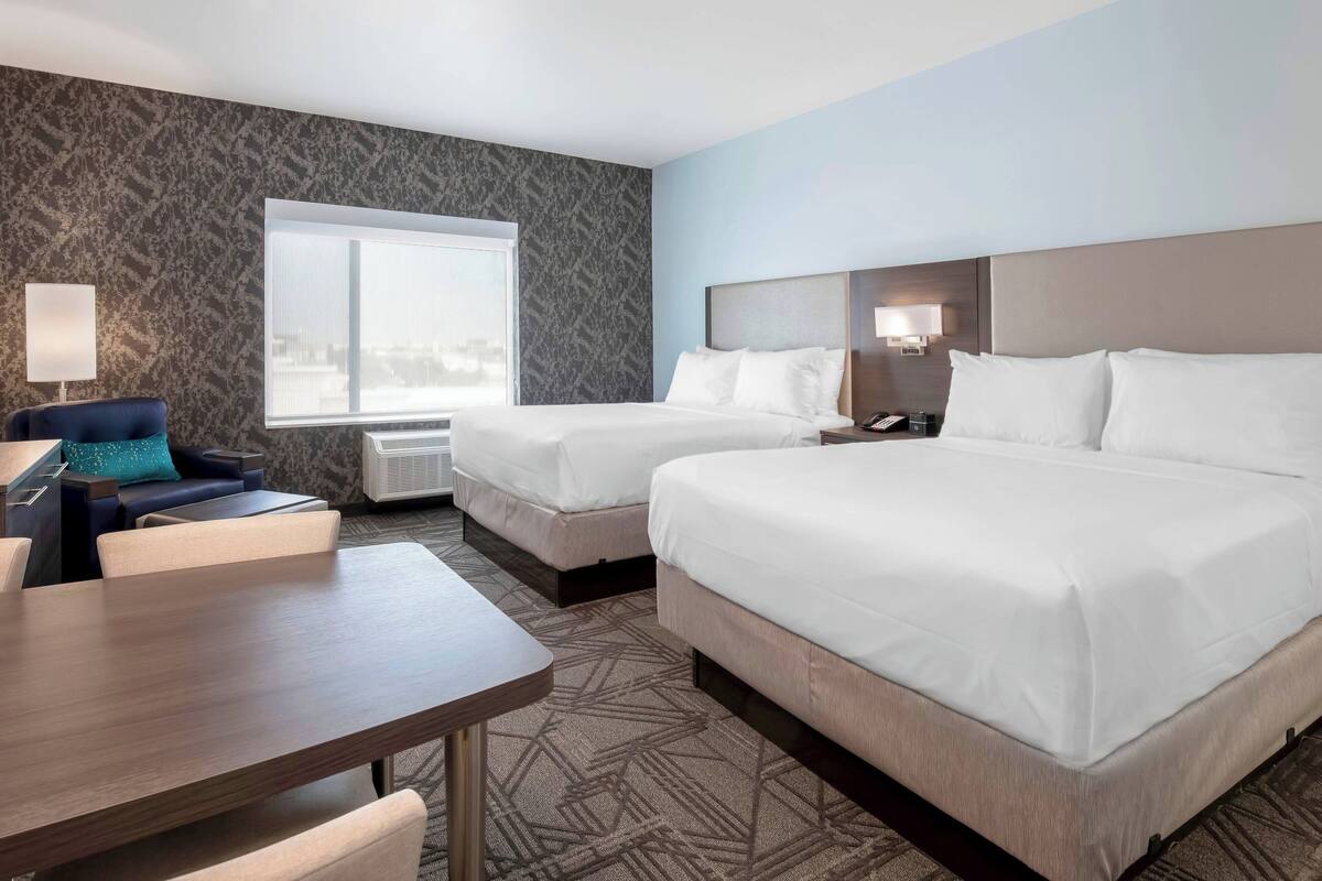 Habitación de hotel con dos camas, un escritorio, una silla, una lámpara y ventana con vista a la ciudad.