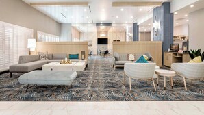 A modern hotel lobby with a central seating area, a dining table, and a TV screen.