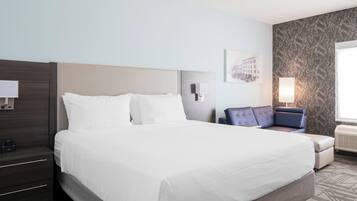 A hotel room with a large bed, a nightstand with a lamp, a seating area with a chair, and a wall-mounted picture.