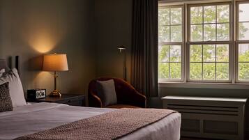A hotel room with a bed, a desk, a chair, a lamp, and a window with a view of trees.
