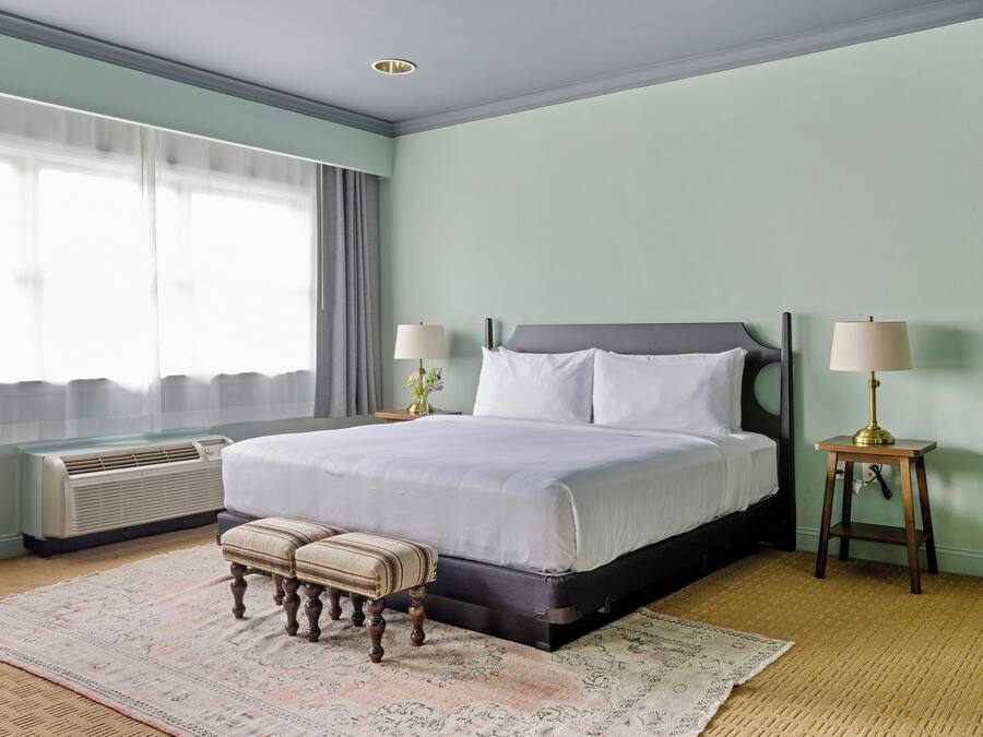 Habitación de hotel con una cama, dos mesitas de noche con lámparas, una ventana con cortinas, un equipo de aire acondicionado y una alfombra estampada.