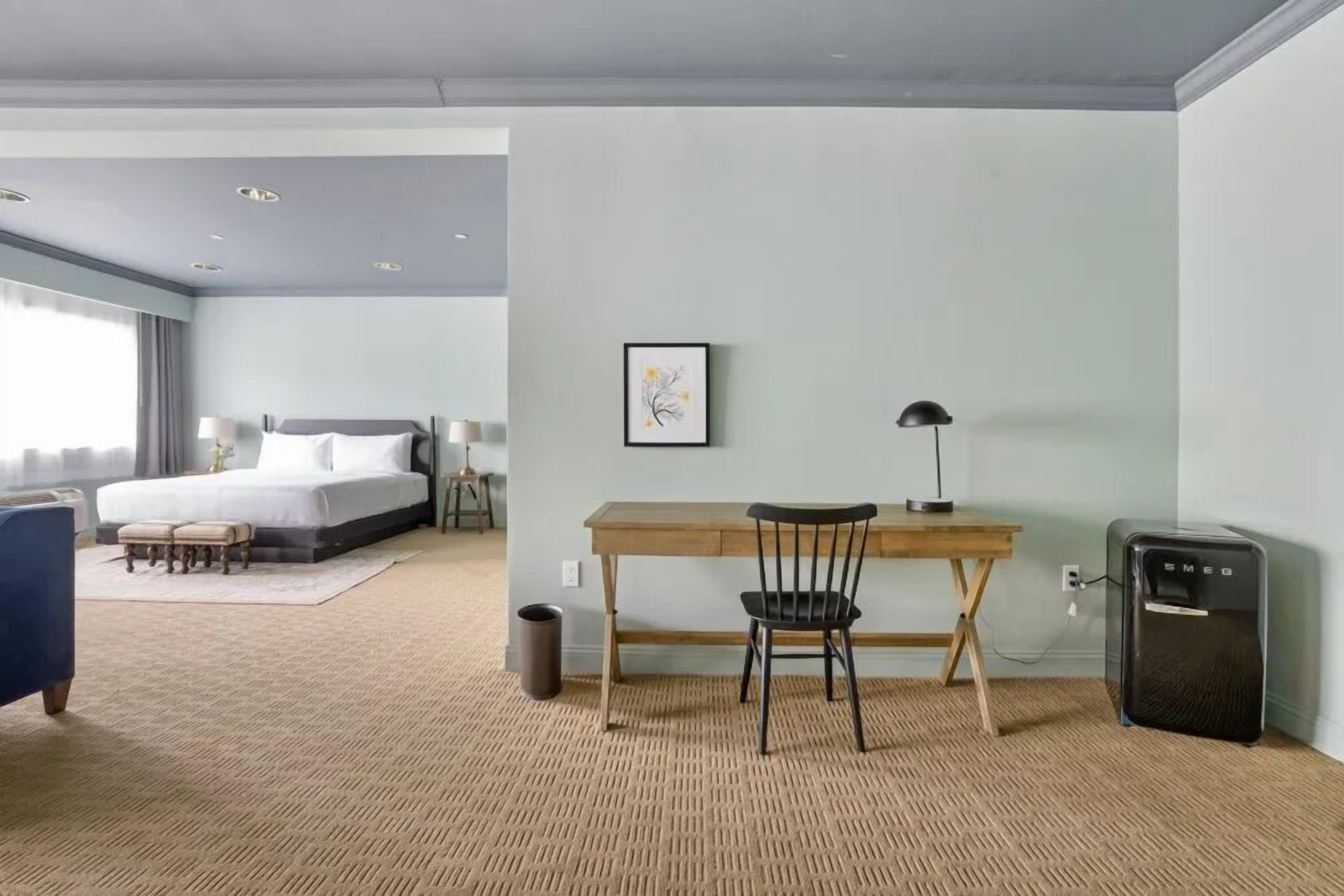 A hotel room with a bed, desk, chair, and a small refrigerator.