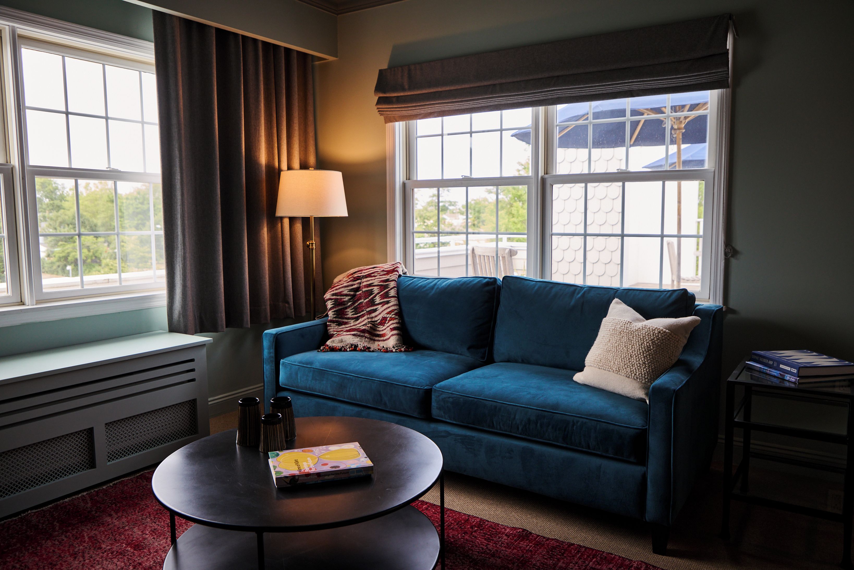 A room with a blue sofa, a round table, a lamp, and a view through large windows.