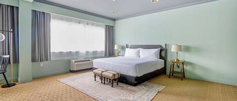 Una habitación de hotel con una cama grande, dos mesitas de noche, una ventana con cortinas, una lámpara de pie y una alfombra estampada.