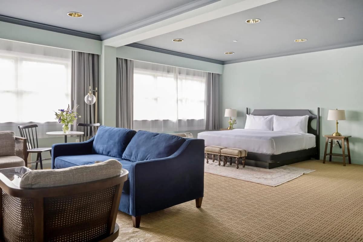 Habitación de hotel con una cama, un sofá azul, una silla de mimbre, una mesita con un florero y una lámpara.