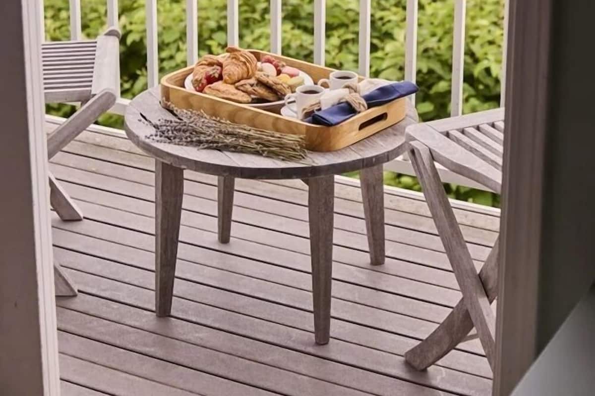 Un balcón con vista a un muelle y un bote, una mesa con una cesta de pasteles, y una silla.