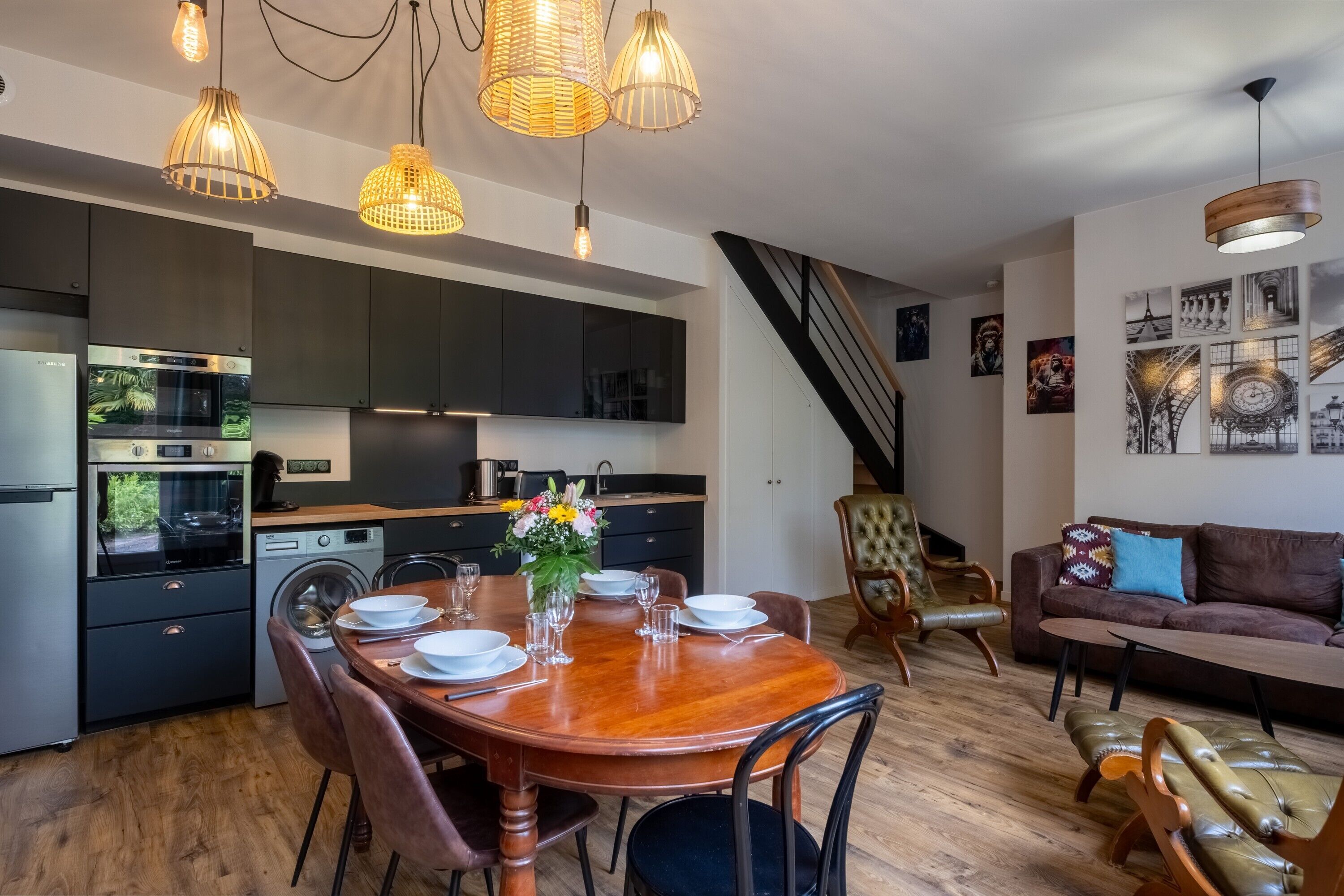 A modern kitchen with dark cabinets, a dining table set for four, and a staircase leading to an upper level.