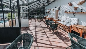 Un patio soleado con sillas de mimbre, un banco de madera y una zona acristalada con mesas y sillas.