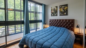 Un dormitorio con un ventanal grande, una colcha azul acolchada, un cabecero marrón con capitonado y dos cuadros en la pared.
