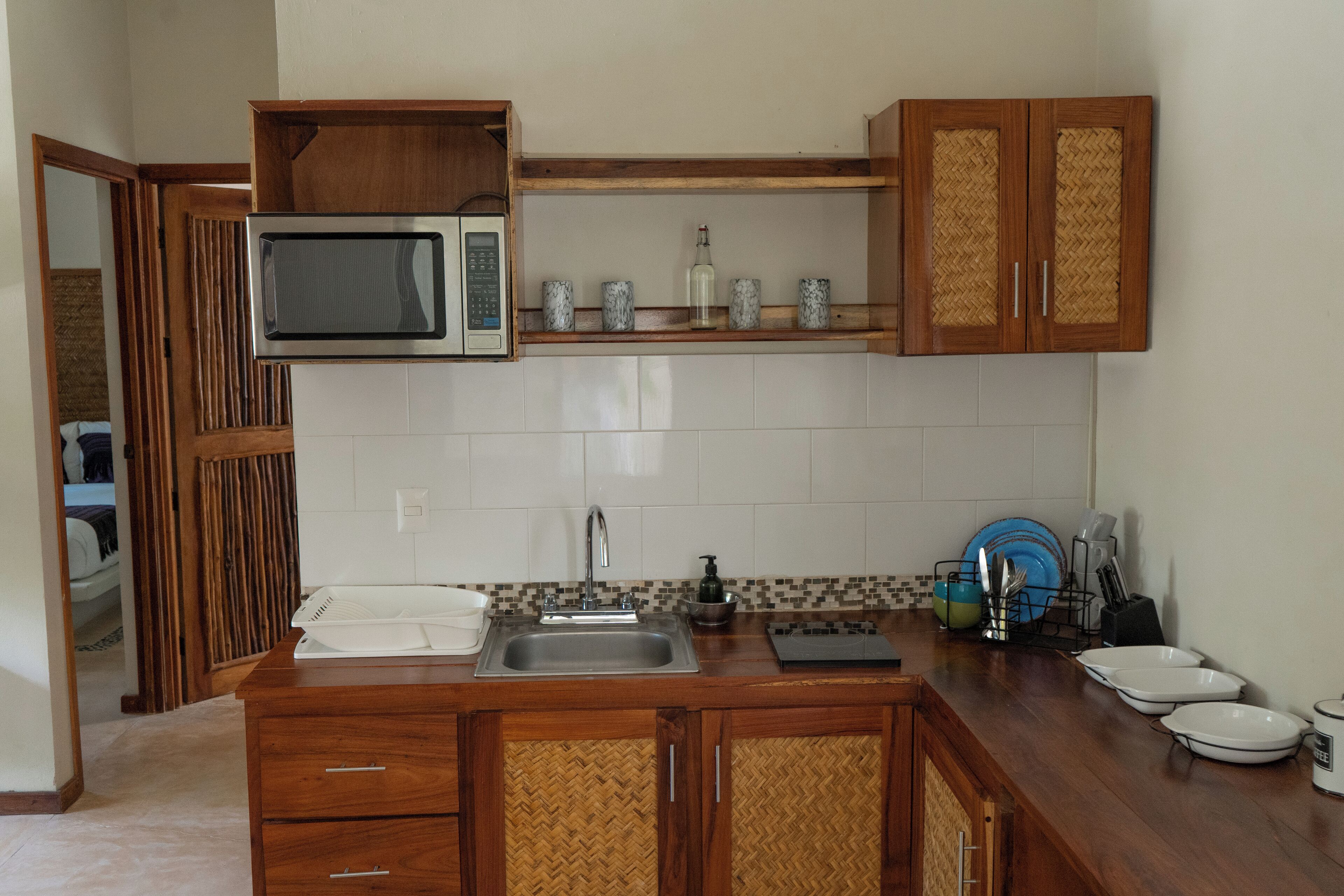 Una cocina con microondas, pileta, armarios de madera y salpicadero de baldosas tipo espiga.