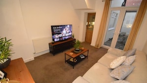 Ein Wohnzimmer mit einer Couch, einem Couchtisch, einem Fernseher, der 'FRIENDS' zeigt, und einer Tür, die auf einen Balkon führt.