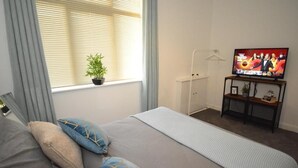 Ein Schlafzimmer mit einem Bett, einem Fernseher, einem Fenster mit Jalousien, einer Pflanze und einem Nachttisch.