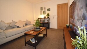 Ein Wohnzimmer mit einer Couch, einem Couchtisch und einem Fernseher. Im Hintergrund sind Wanddekorationen und eine Tür zu sehen.