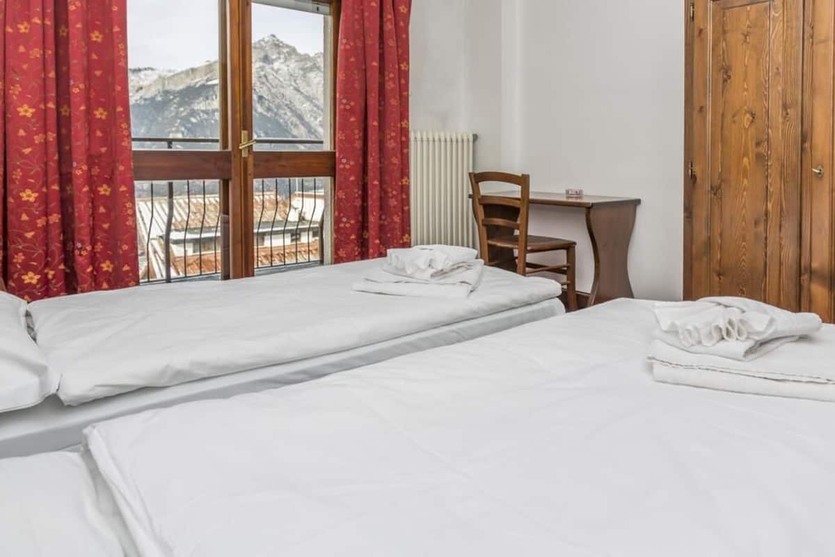 A hotel room with two beds, a wooden chair, a small table, and a view of mountains through a window with red curtains.